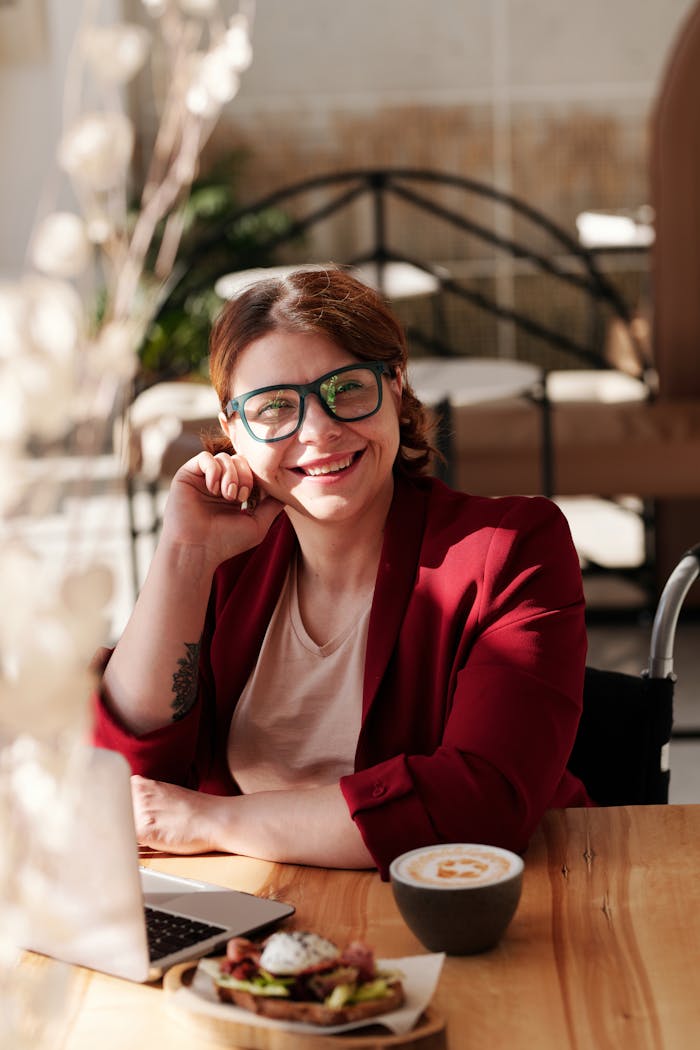 Smiling woman in red blazer working on laptop with coffee and meal at café.